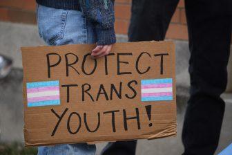 Archival photo of an individual holding a protest sign with a transgender flag. March 12, 2021.