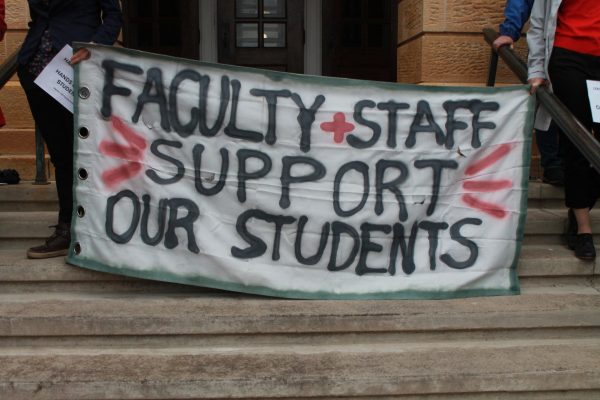 Faculty protest outside of Bascom Hall. Two faculty hold this sign reading FACULTY + STAFF SUPPORT OUR STUDENTS. May 2, 2024.