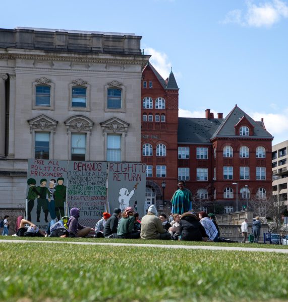 People sit on the grass at an SJP teach-in. The sign behind them reads "Free all political prisoners," and "DEMANDS: 1) Disclosure of UW foundation's investments. 2) Divest from prisons, borders & occupation. 3) Fight visa revocation, no UW-ICE cooperation."